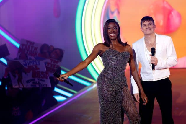 Two smiling TV hosts on a colorful stage holding microphones, with cheering fans and signs in the background